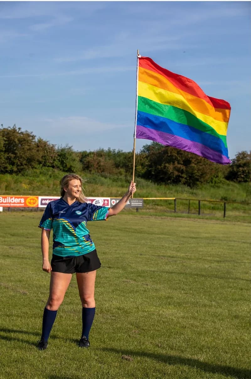 Sarah Lynch standing on the rugby pitch holding a Pride flag
